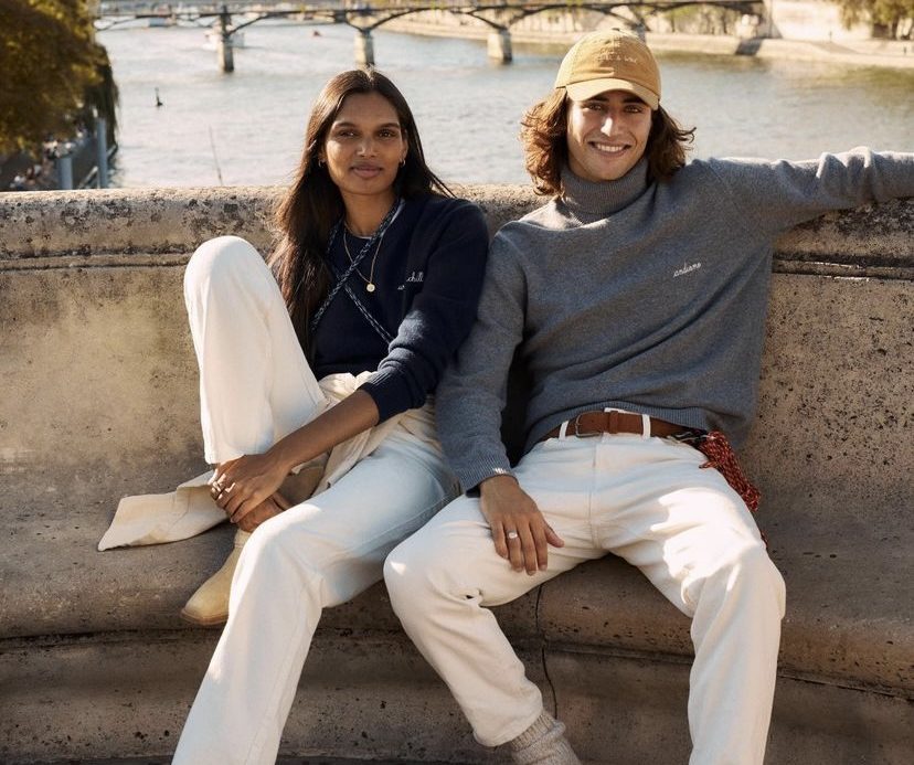 Auf dem Bild sitzen eine modisch gekleidete Frau und ein Mann lässig auf einer Bank am Hafen. Beide tragen Kleidung von Maison Labiche und strahlen eine entspannte Atmosphäre aus.