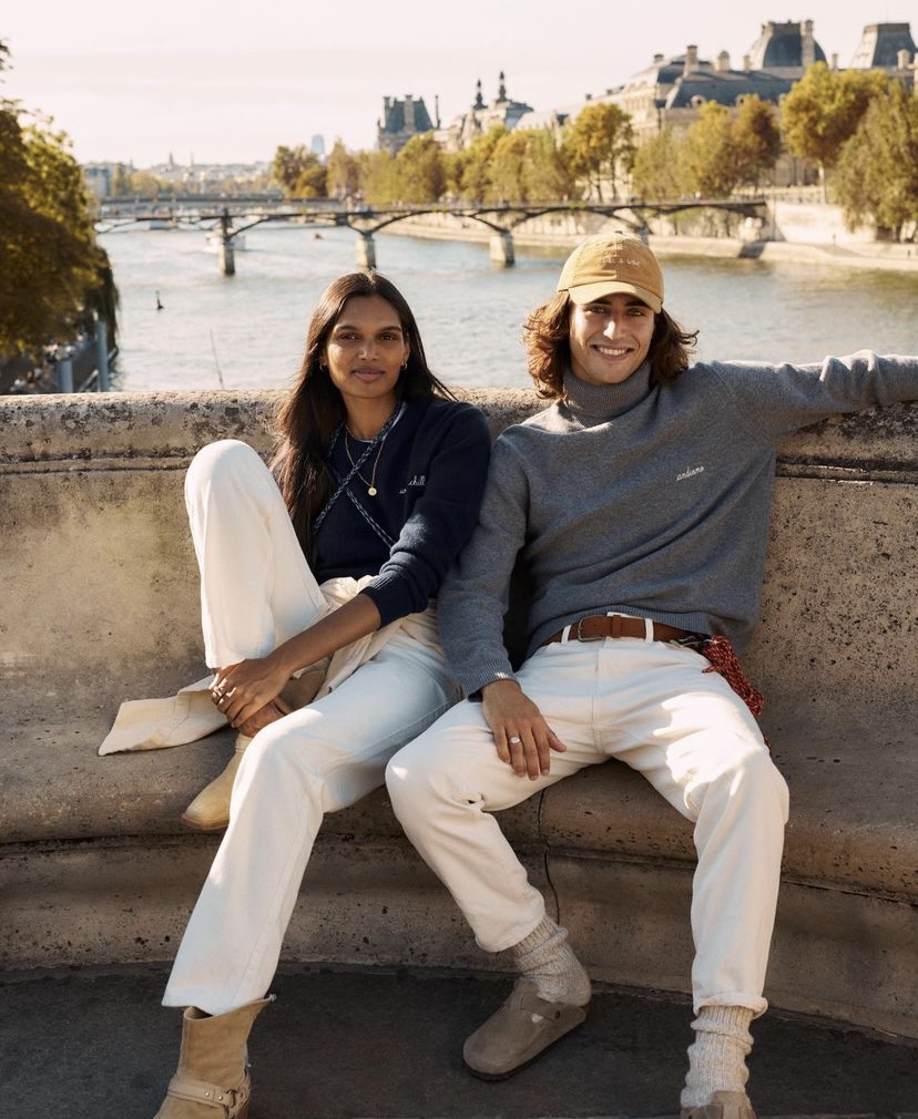 Auf dem Bild sitzen eine modisch gekleidete Frau und ein Mann lässig auf einer Bank am Hafen. Beide tragen Kleidung von Maison Labiche und strahlen eine entspannte Atmosphäre aus.